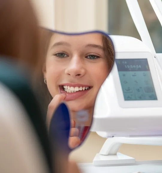 Usmívající se dívka si prohlíží zuby v zrcátku v zubařském křesle vedle dentální soupravy na stomatologii AGEL PLUS
